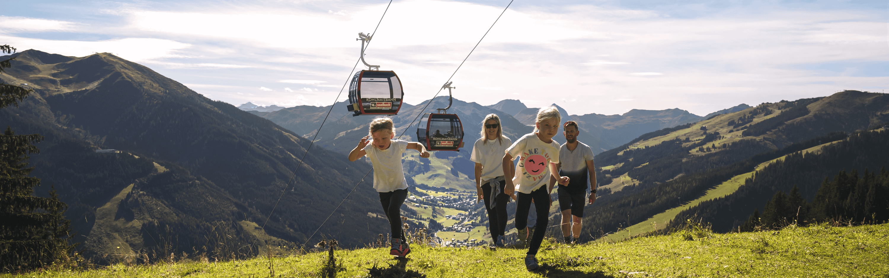 En familie med to børn går op ad en græsklædt bakke under en svævebanelinje. Bjerget landskab med en dal og by i baggrunden. Himlen er klar og solrig, hvilket skaber en malerisk udendørs scene.