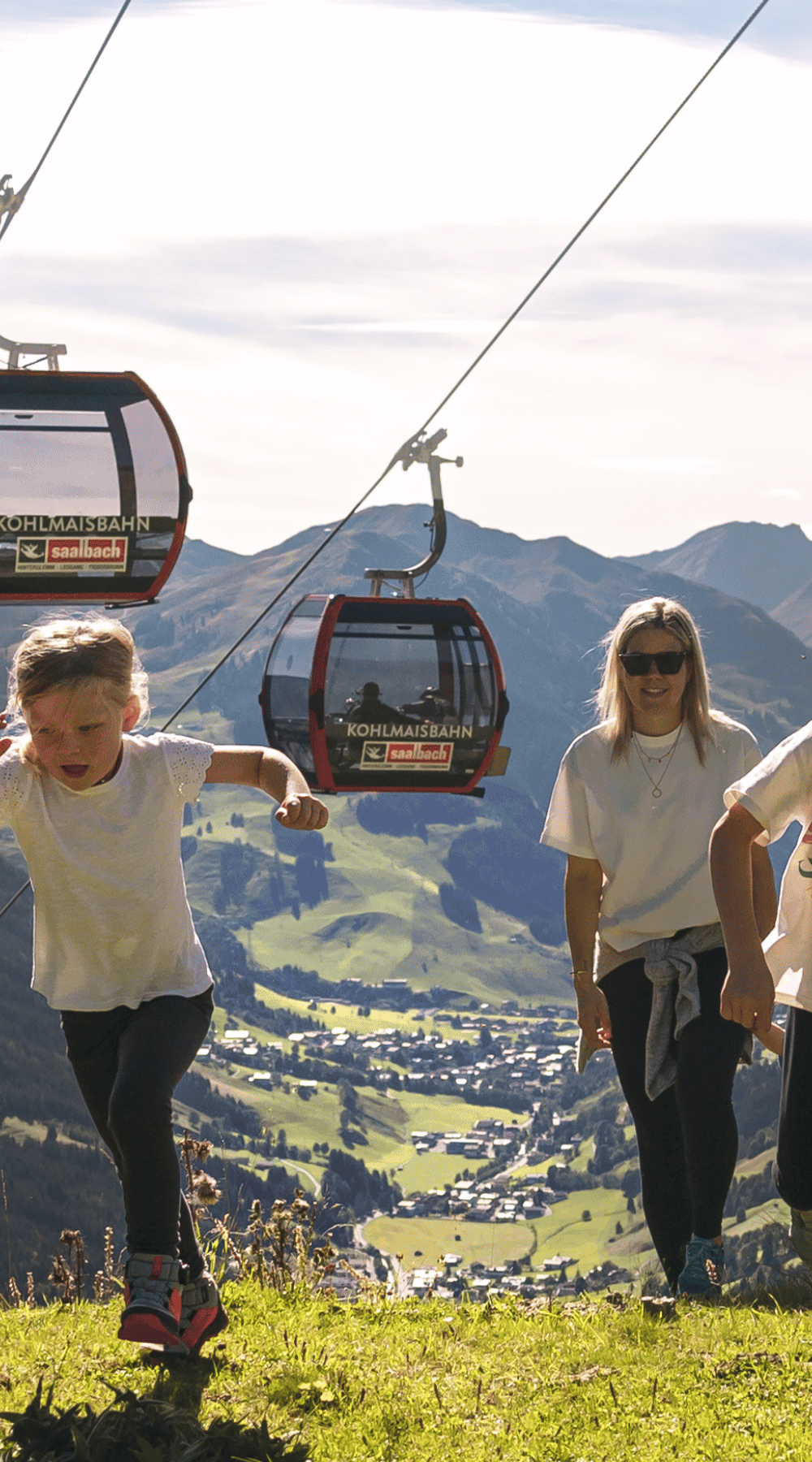 En familie med to børn går op ad en græsklædt bakke under en svævebanelinje. Bjerget landskab med en dal og by i baggrunden. Himlen er klar og solrig, hvilket skaber en malerisk udendørs scene.