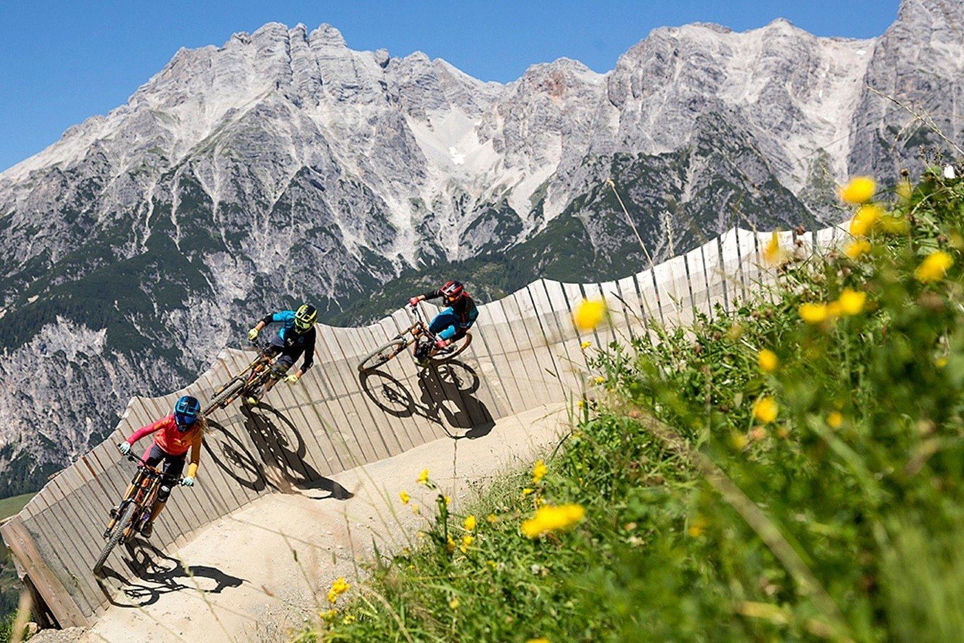 Tre cyklister cyklar mountainbikes på en kurvig träbana omgiven av grönt gräs och gula blommor, med Wagrains karga berg i bakgrunden under en klarblå himmel – ett perfekt skidresmål på sommaren.