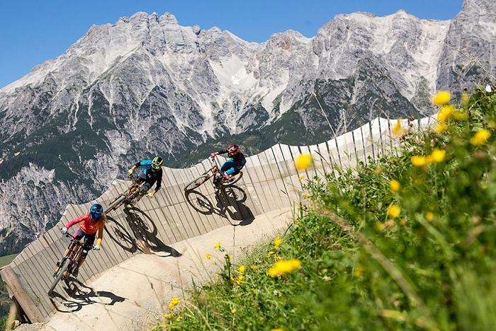 Tre cyklister cyklar mountainbikes på en kurvig träbana omgiven av grönt gräs och gula blommor, med Wagrains karga berg i bakgrunden under en klarblå himmel – ett perfekt skidresmål på sommaren.