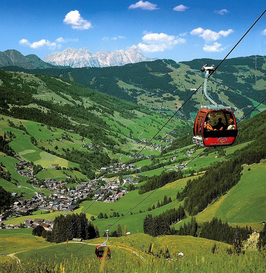 En rød svævebane stiger op over en frodig grøn dal i Sommerferie i Saalbach, med en lille landsby beliggende nedenfor. Bølgende bakker og tætte træer omgiver området, mens fjerne bjerge skaber en naturskøn kulisse under en klar blå himmel.