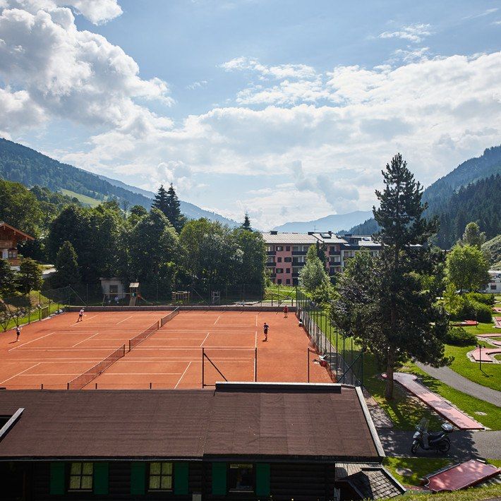 Midt i en naturskøn udsigt over en tennisbane omgivet af frodige grønne træer og bjerge under en delvist overskyet himmel, nyder spillerne den orange lerbane. Denne idylliske scene, der minder om en sommerferie i Saalbach, er indrammet af bygninger og blomstrende grønne områder i baggrunden.