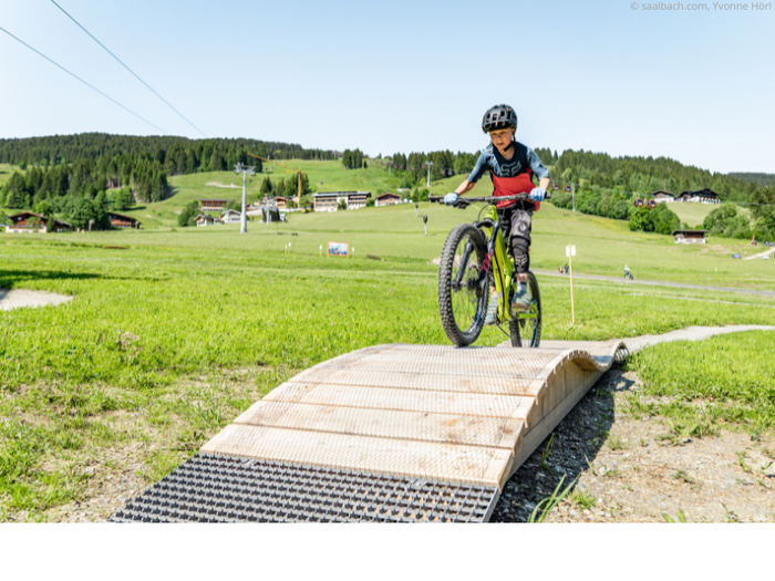 Ett litet barn med hjälm cyklar mountainbike uppför en liten ramp på ett gräsbevuxet fält i Wagrain, med kullar, träd och byggnader i bakgrunden – perfekt för en aktiv sommarferie i Österrike en solig dag.