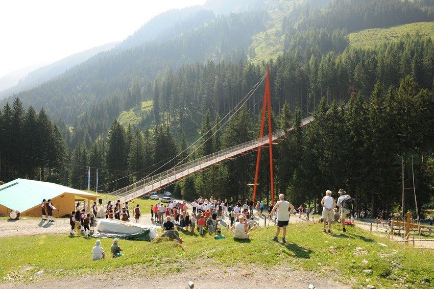 Under sommerferien i Saalbach samledes en menneskemængde ved en lang hængebro omgivet af frodige grønne bakker og tætte træer. En lille bygning med grønt tag er synlig, og scenen er sat på en solskinsdag i dette maleriske bjerglandskab.