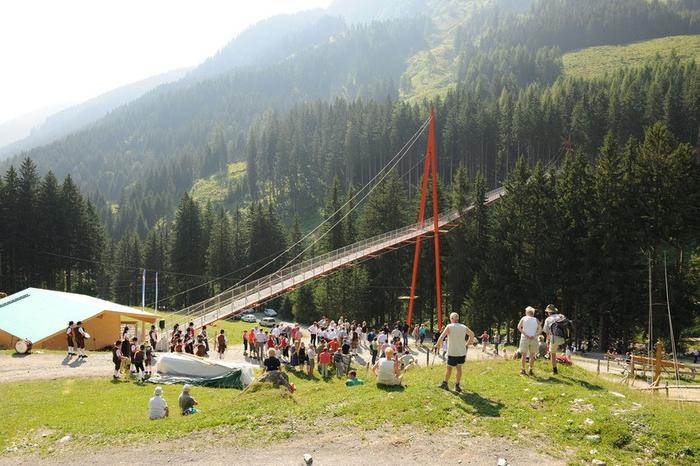 Under sommerferien i Saalbach samledes en menneskemængde ved en lang hængebro omgivet af frodige grønne bakker og tætte træer. En lille bygning med grønt tag er synlig, og scenen er sat på en solskinsdag i dette maleriske bjerglandskab.