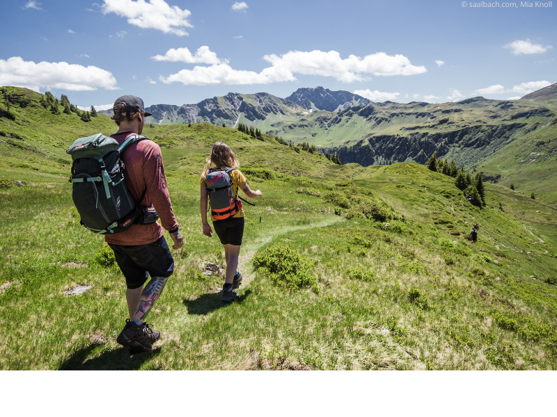To vandrere med rygsække går ad en grøn bjergsti under en lys himmel i Saalbach. Bølgende bakker og fjerne tinder omgiver dem, mens solen kaster skygger på det frodige landskab, der minder om stier nær Westendorf, der er berømt for skirejseeventyr.