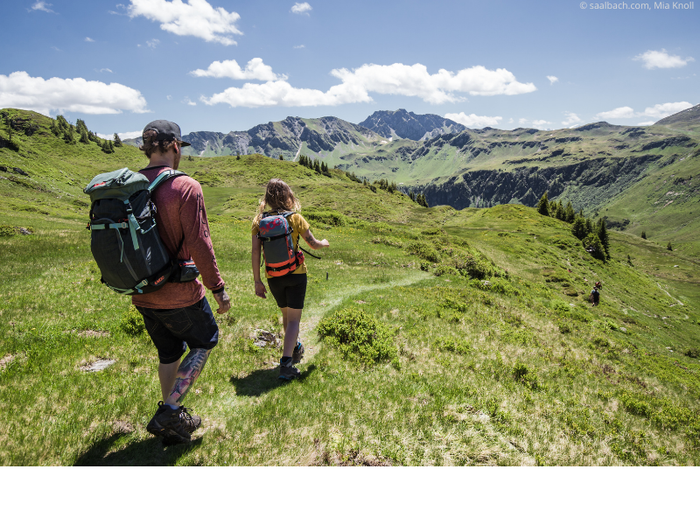 To vandrere med rygsække går ad en grøn bjergsti under en lys himmel i Saalbach. Bølgende bakker og fjerne tinder omgiver dem, mens solen kaster skygger på det frodige landskab, der minder om stier nær Westendorf, der er berømt for skirejseeventyr.