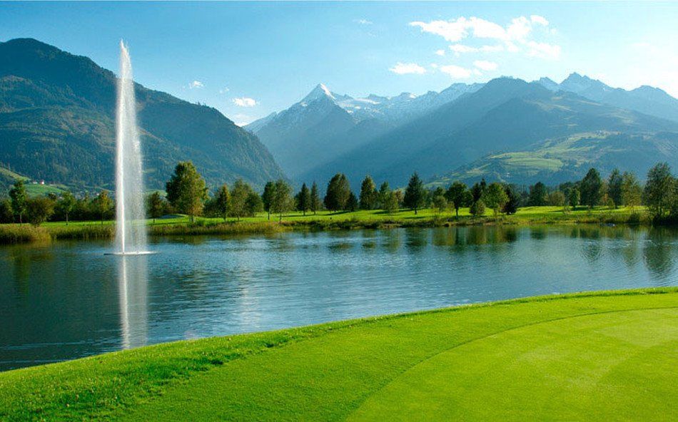 En golfbana med grön gräsmatta gränsar till en lugn sjö med en hög vattenfontän, omgiven av träd och berg under en solig, klarblå himmel – perfekt för att kombinera avkoppling med skidresor till Wagrain eller Westendorf.