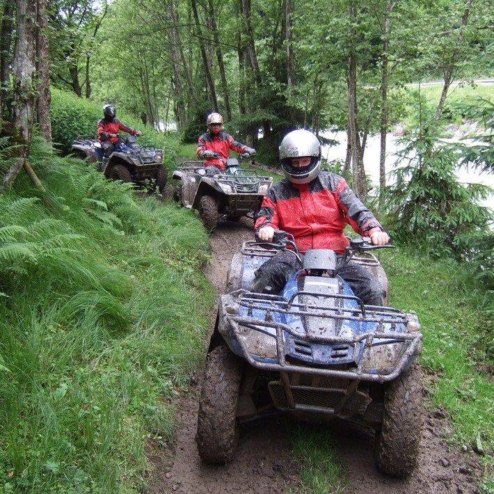 Tre personer i hjelme og røde jakker kører i terrængående køretøjer (ATV'er) på en mudret skovsti og oplever spændingen ved en Sommerferie i Saalbach. Frodigt grønt løv omgiver stien med høje træer og bregner synlige. En flod flyder i baggrunden under en overskyet himmel.