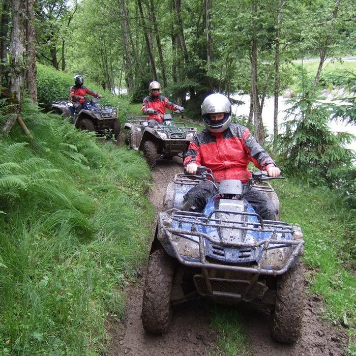 Tre personer i hjelme og røde jakker kører i terrængående køretøjer (ATV'er) på en mudret skovsti og oplever spændingen ved en Sommerferie i Saalbach. Frodigt grønt løv omgiver stien med høje træer og bregner synlige. En flod flyder i baggrunden under en overskyet himmel.
