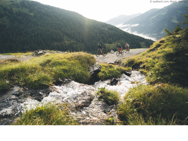 Tre cyklister cyklar mountainbikes längs en grusväg i Saalbachs frodiga, gröna alpina landskap, med en liten bäck som rinner i förgrunden och skogsklädda kullar och dimmiga berg – perfekt för sommerferie Österrig – som reser sig i bakgrunden.