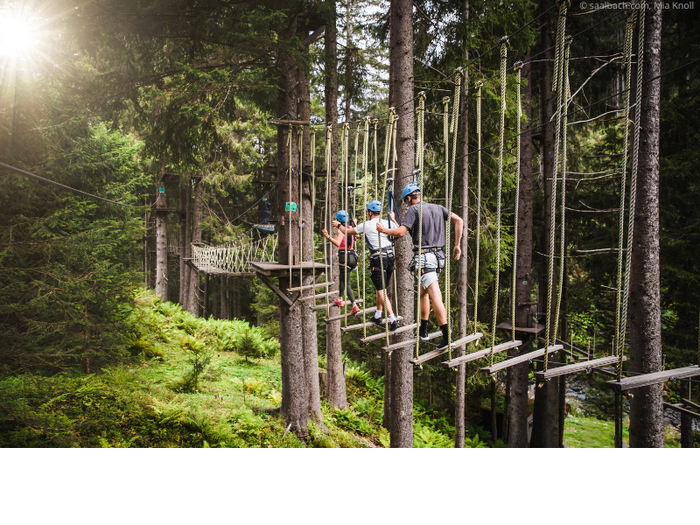 Tre personer i hjälmar och selar går på upphängda träplankor och rep i en skogsäventyrspark i Saalbach, omgivna av höga träd och solljus som silar genom grenarna. Perfekt för dig som söker spänning under sommarferien i Österrike.