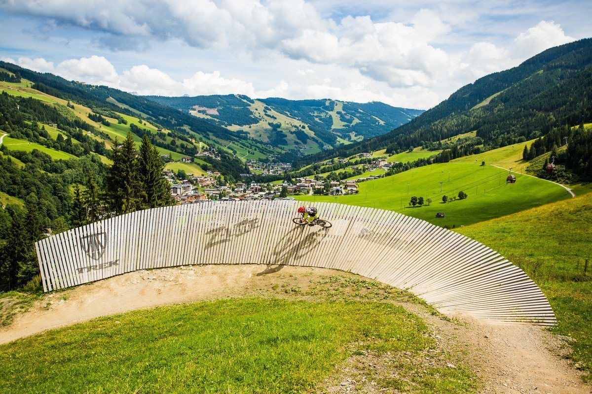 En mountainbiker navigerer på en buet trævæg på en græsbakke under sommerferie i Saalbach, med en naturskøn udsigt over en frodig dal og spredte bygninger i baggrunden under en delvist overskyet himmel.