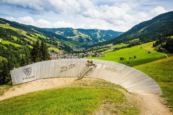 En mountainbiker navigerer på en buet trævæg på en græsbakke under sommerferie i Saalbach, med en naturskøn udsigt over en frodig dal og spredte bygninger i baggrunden under en delvist overskyet himmel.