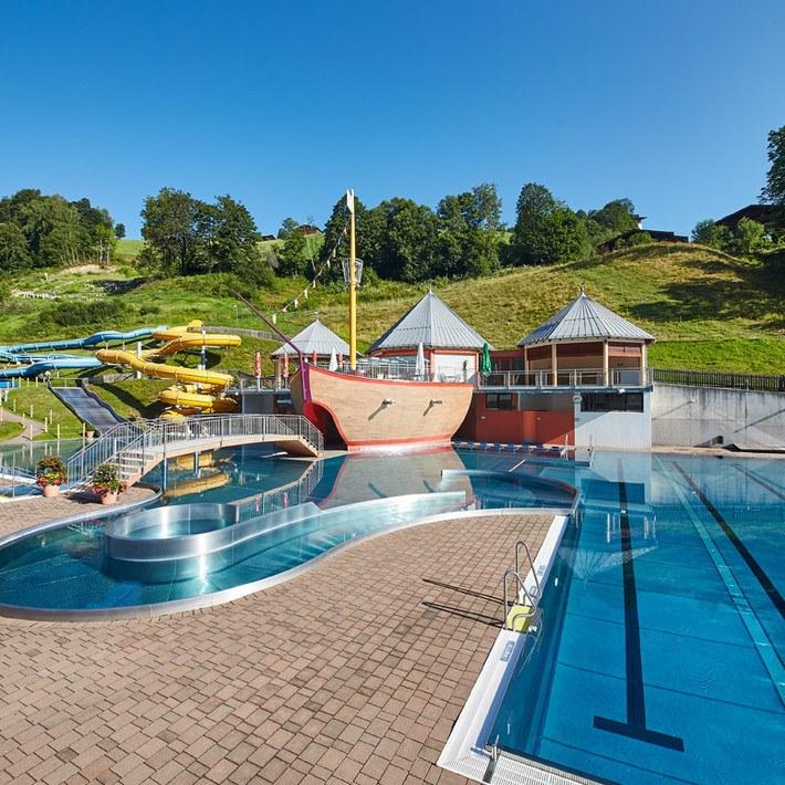 Oplev et stænk af sjov under din Sommerferie i Saalbach på vores udendørs svømmeområde, med en stor pool, skibstema legestruktur og farverige vandrutsjebaner. Omgivet af frodige grønne områder og træer under en klar blå himmel, er det den perfekte sommerferie.