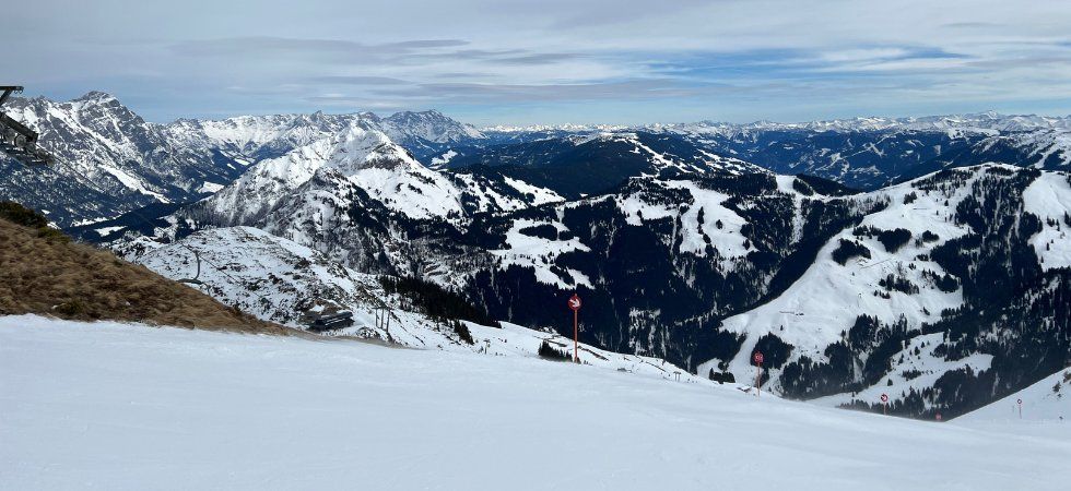 Snötäckta berg under en delvis molnig himmel, med skidbackar och röda skidmarkörer i förgrunden - perfekt för en skiferie i Saalbach. Fjärran toppar och dalar sträcker sig in i bakgrunden.