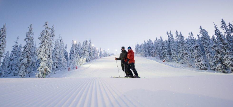 Två skidåkare står tillsammans i en nypreparerad snöig backe, omgiven av snötäckta träd under en klar himmel i gryning eller skymning - ett perfekt ögonblick på en skiferie i Sälen, lugnt och vintrigt.