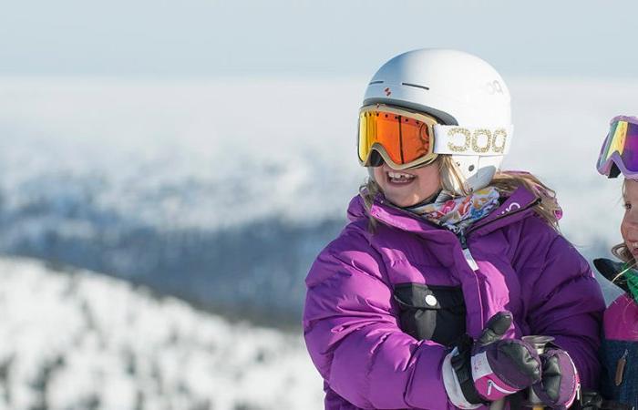 Två barn med skidhjälm, skidglasögon och vinterjacka ler och står tillsammans utomhus med ett snöigt fjällandskap i bakgrunden - en bild som fångar glädjen i ett skiferie i Sälen.