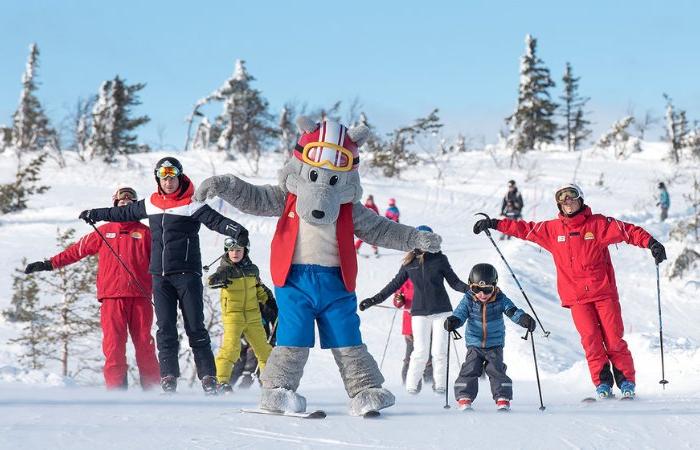 En grupp människor och barn åker skidor i en snöig backe under sitt skiferie i Sälen, ledda av en person i en stor, grå djurmaskotdräkt med skyddsglasögon, väst och shorts. I bakgrunden syns snötäckta träd och en klar himmel.