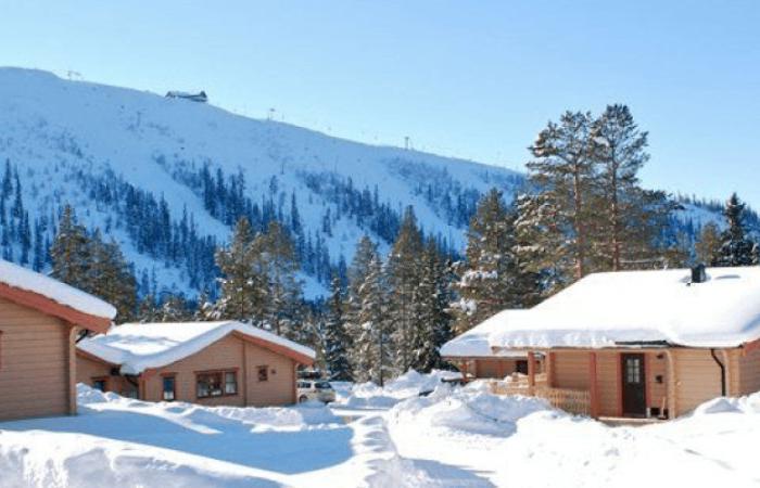 Trästugor med snötäckta tak omgivna av träd, mot en snöig fjällbakgrund under en klarblå himmel - perfekt för en avkopplande skiferie i Sälen.