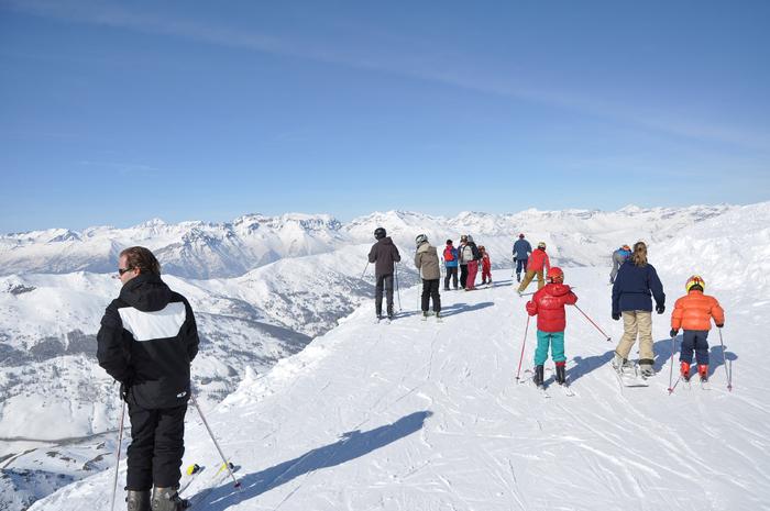 En grupp människor, utrustade med skidutrustning, står på toppen av en snöig bergstopp i Westendorf. De har utsikt över en stor, snötäckt bergskedja under en klarblå himmel, och förbereder sig för att åka skidor nerför backen.