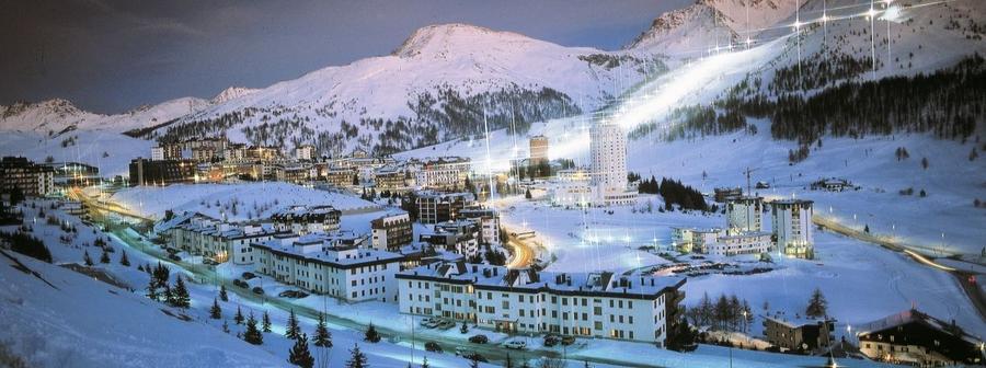 En sneklædt bjerglandsby i skumringen, der minder om skiferie i Westendorf, med oplyste bygninger og gader. Landskabet er omgivet af bjerge under en klar, blå himmel. Det snedækkede område skaber en fredfyldt vinterstemning.