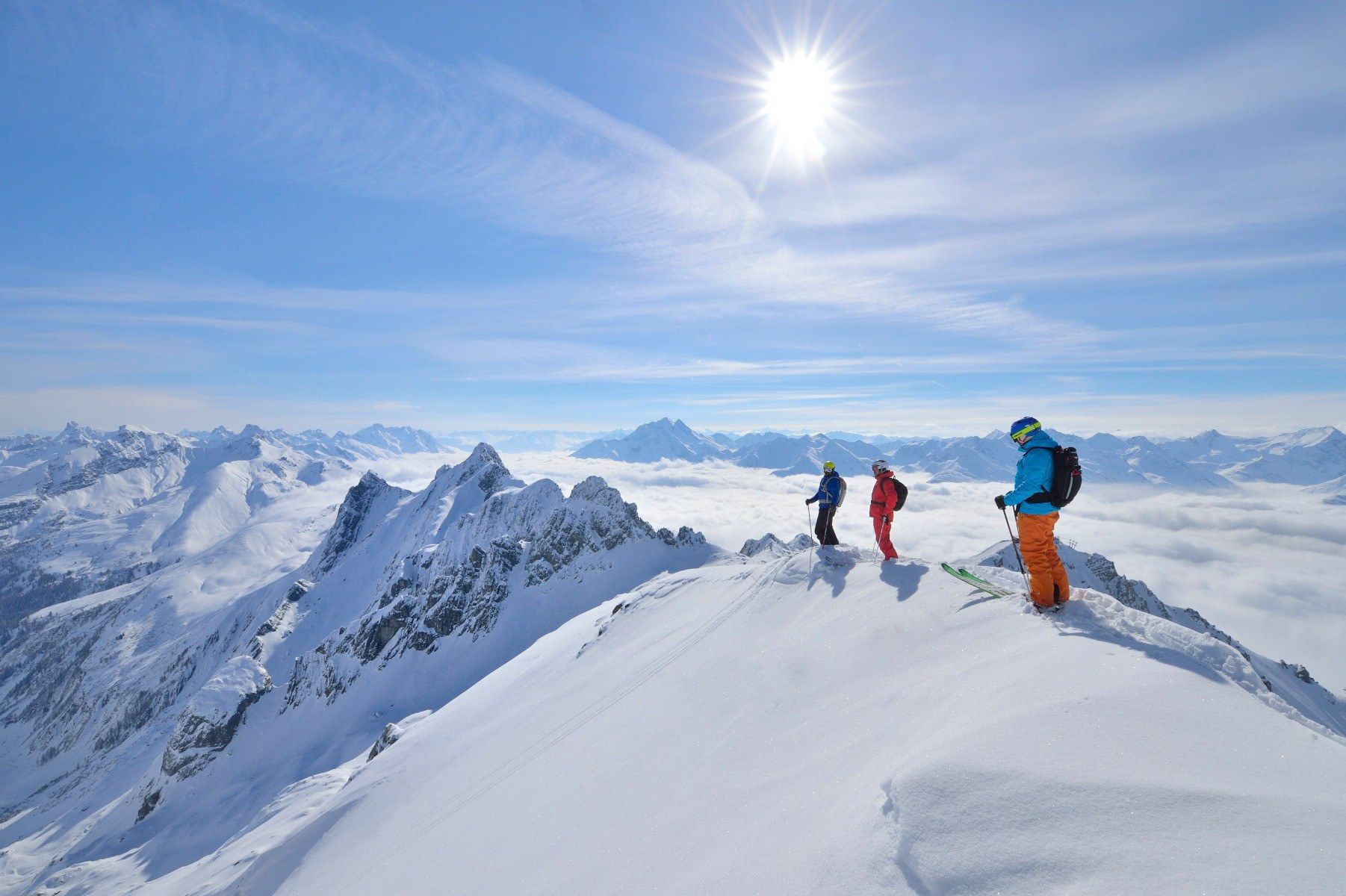 Tre skidåkare står på toppen av en snöig bergskam under en strålande sol, omgivna av snötäckta toppar nära Saalbach och ett molnhav i fjärran, och förbereder sig för att påbörja sin oförglömliga skidåkningstur.