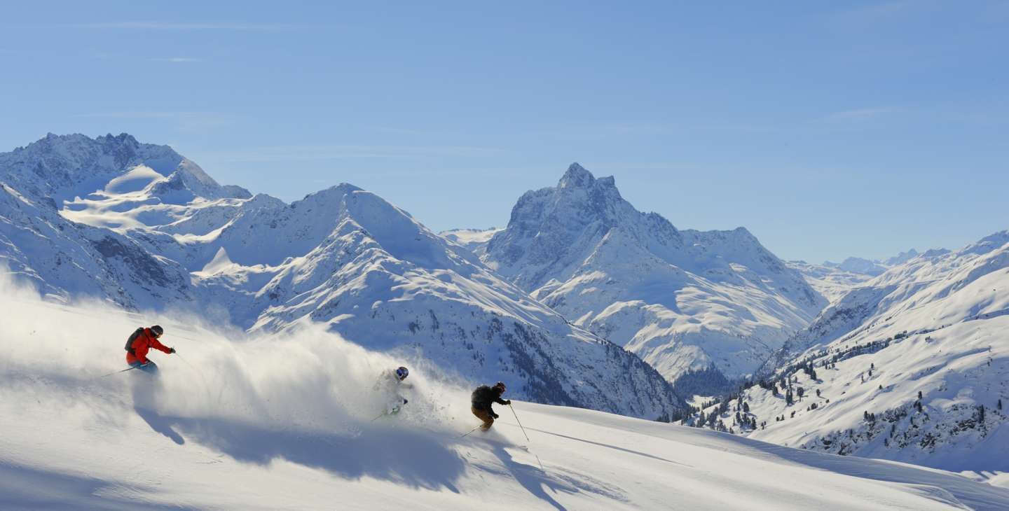 Tre skiløbere går ned ad en sneklædt bjergskråning, omgivet af majestætiske tinder under en klar blå himmel i hjertet af en skiferie i St. Anton. Pudderagtige snestier bag dem, hvilket skaber en dynamisk scene for vintersport.