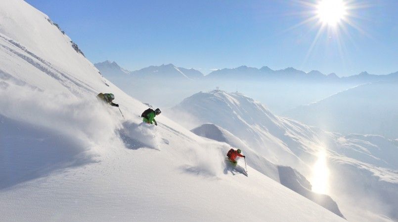 Tre skidåkare i färgglada kläder tar sig ner för en snöig bergssluttning i Wagrain under en klarblå himmel, med solen skinande starkt. I bakgrunden syns bergstoppar genom ett lager av dimma.