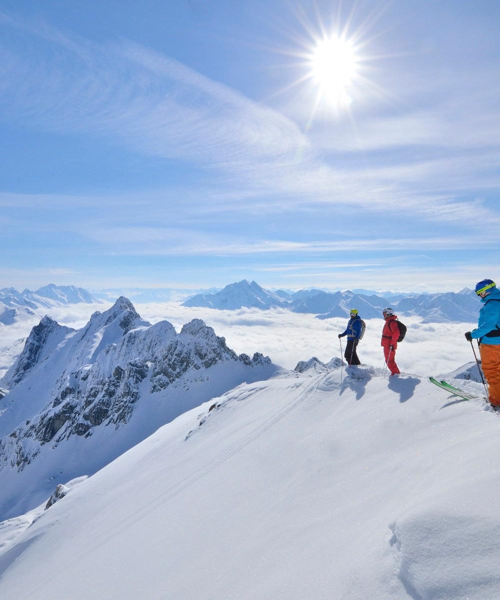 Tre skiløbere i farverigt udstyr står på en sneklædt bjergryg under deres skiferie i St. Anton. Den klare sol og den klare blå himmel oplyser store snedækkede tinder og skyer, der strækker sig ud i baggrunden og skaber et betagende alpint panorama.
