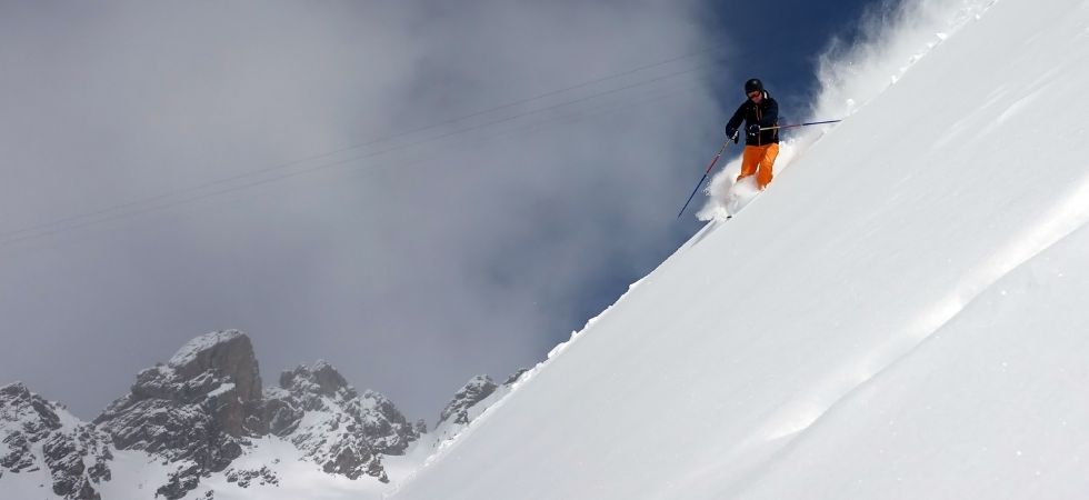 En skidåkare i orange byxor åker nerför en brant, snöig bergssluttning på en skiferie i St. Anton, omgiven av puder med robusta, snötäckta toppar i bakgrunden under en delvis molnig himmel.