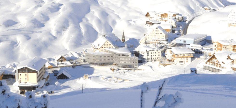 En liten bergsby i St Anton är omgiven av snötäckta kullar och byggnader med snötäckta tak i starkt solljus - perfekt för en skiferie i St Anton.