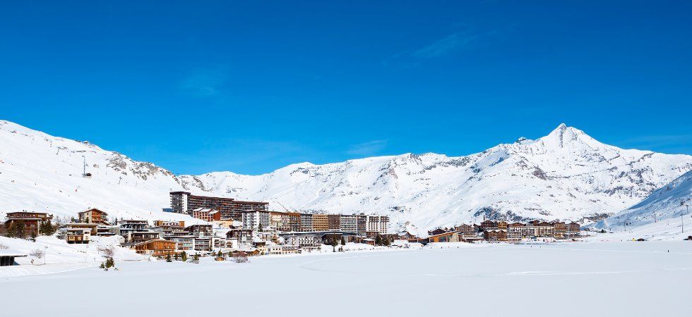 En snöig bergsort med byggnader som ligger samlade vid foten av höga, snötäckta toppar, under en klarblå himmel - perfekt för en skiferie i Tignes. I förgrunden syns en vidsträckt, snötäckt slätt.