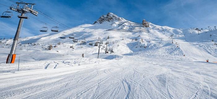 Snötäckt skidbacke med stolliftar till vänster, som leder upp på ett berg under en klarblå himmel. Skidspår markerar den lugna och ljusa scenen - perfekt för en skiferie i Tignes.