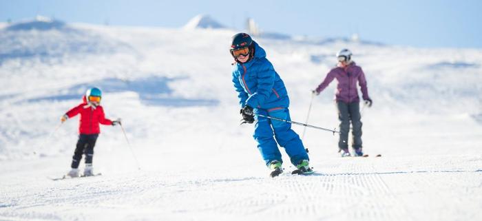 Tre personer njuter av en skiferie i Trysil, skidåkning på en snötäckt sluttning under en klarblå himmel. Ett leende barn i blå utrustning står i förgrunden och två andra följer efter i röda och lila jackor.