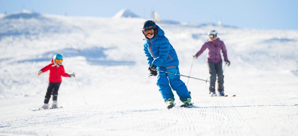 Tre personer njuter av en skiferie i Trysil, skidåkning på en snötäckt sluttning under en klarblå himmel. Ett leende barn i blå utrustning står i förgrunden och två andra följer efter i röda och lila jackor.