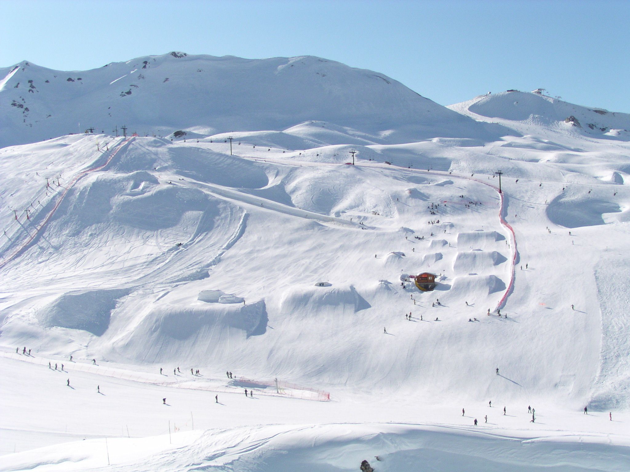 En snörik skidort i Westendorf med svagt sluttande kullar och några branta toppar. Skid- och snowboardåkare navigerar i olika nedfarter och hopp. En skidlift och en liten byggnad är synliga, mot en klarblå himmel, perfekt för minnesvärda skidresor.