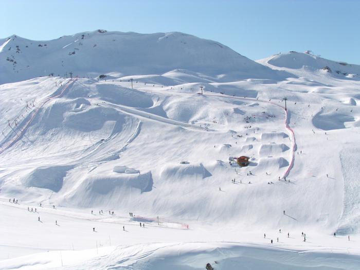 En snörik skidort i Westendorf med svagt sluttande kullar och några branta toppar. Skid- och snowboardåkare navigerar i olika nedfarter och hopp. En skidlift och en liten byggnad är synliga, mot en klarblå himmel, perfekt för minnesvärda skidresor.