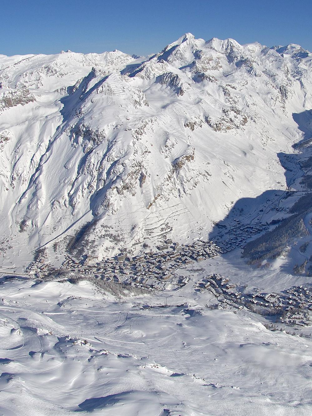 Flygfoto över det snötäckta bergslandskapet nära Saalbach, med en by inbäddad i dalen. Topparna skimrar under sin snöiga filt, och den klarblå himlen förstärker den krispiga, kalla atmosfären. Byn har spridda byggnader och slingrande vägar.