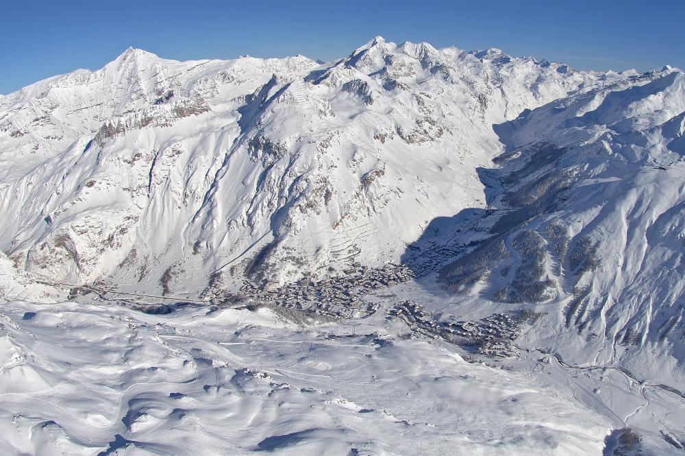 Flygfoto över det snötäckta bergslandskapet nära Saalbach, med en by inbäddad i dalen. Topparna skimrar under sin snöiga filt, och den klarblå himlen förstärker den krispiga, kalla atmosfären. Byn har spridda byggnader och slingrande vägar.