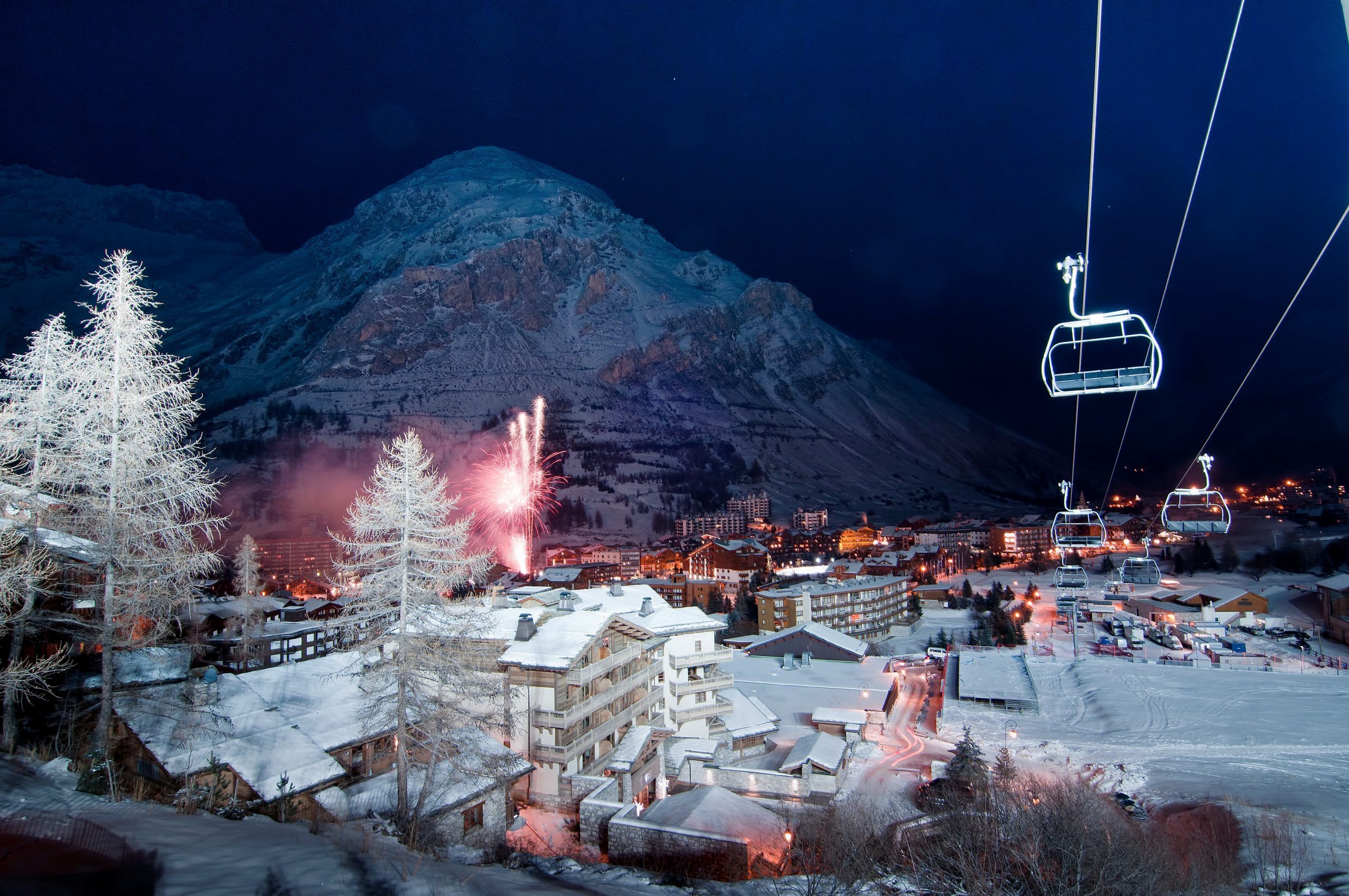 En snötäckt bergsstad på natten, upplyst av fyrverkerier på himlen. Skidliftar hänger över Saalbachs snötäckta landskap, och byggnader med upplysta fönster ligger inbäddade bland snötäckta träd och sluttningar.