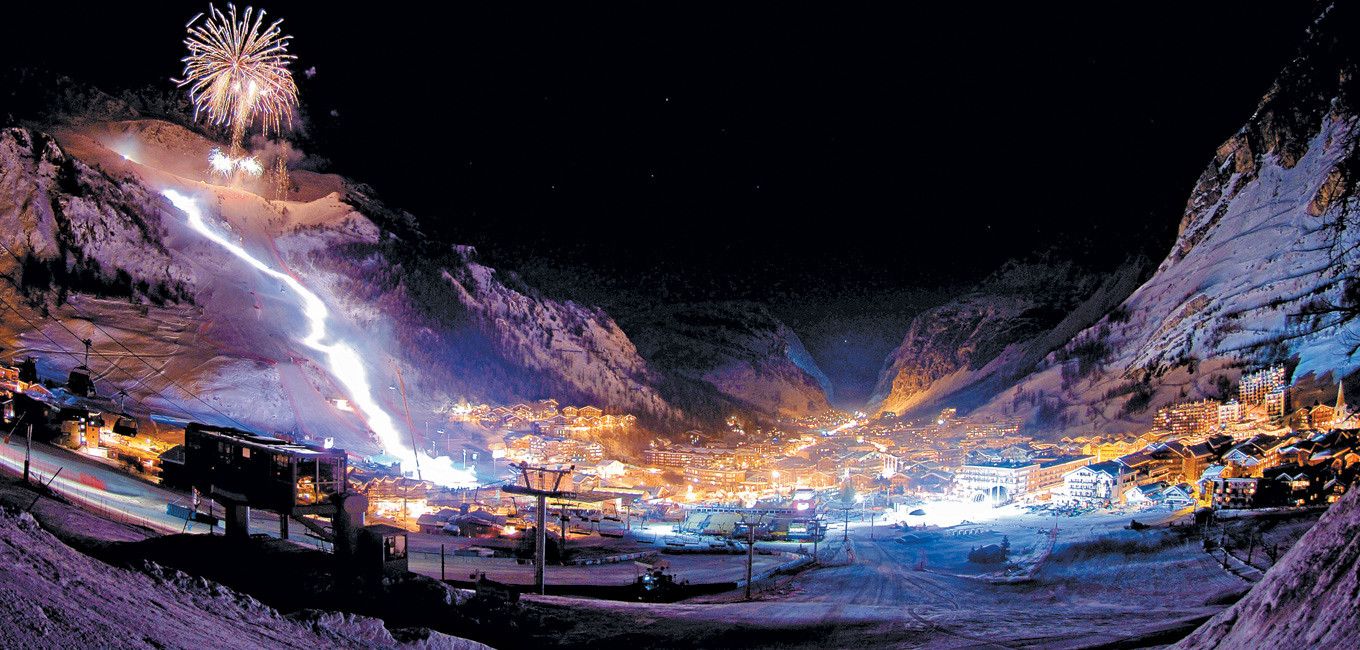 Wagrains snöiga bergsresort lyser på natten, med upplysta skidbackar och starkt upplysta byggnader som skapar ett vinterlandskap. Fyrverkerier målar himlen, allt ställt mot mörka, höga berg under en stjärnklar himmel – en perfekt destination för skidresor eller skidresor.