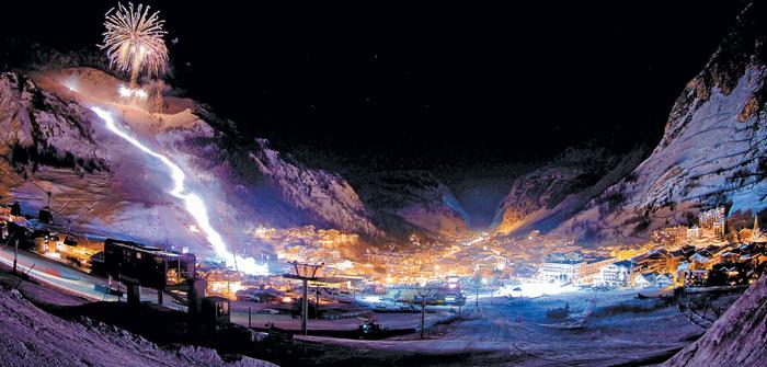 Wagrains snöiga bergsresort lyser på natten, med upplysta skidbackar och starkt upplysta byggnader som skapar ett vinterlandskap. Fyrverkerier målar himlen, allt ställt mot mörka, höga berg under en stjärnklar himmel – en perfekt destination för skidresor eller skidresor.