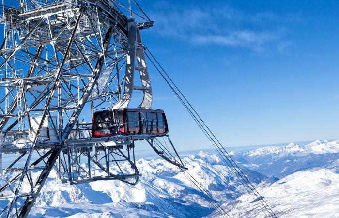 En svævebane afgår fra en metalstation højt i de sneklædte bjerge under en skiferie. Himlen er klar og blå og tilbyder panoramaudsigt over de omkringliggende snedækkede tinder og dale. Nogle få mennesker står på platformen i Wagrain og observerer det betagende landskab.