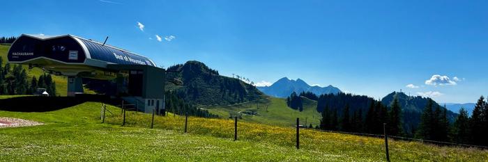 Ett gräsbevuxet bergslandskap med en skidliftstation till vänster, böljande kullar och avlägsna toppar under en klarblå himmel - perfekt för en Sommerferie i Wagrain. Vilda blommor och träd fyller den livfulla förgrunden.