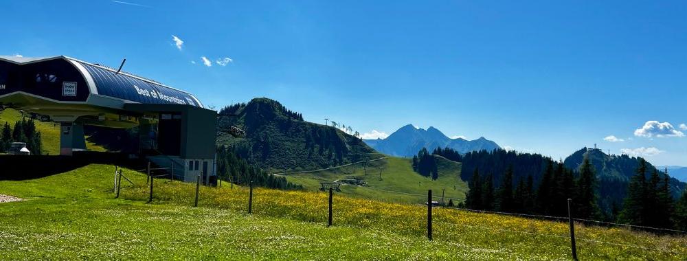 Ett gräsbevuxet bergslandskap med en skidliftstation till vänster, böljande kullar och avlägsna toppar under en klarblå himmel - perfekt för en Sommerferie i Wagrain. Vilda blommor och träd fyller den livfulla förgrunden.