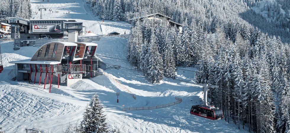 En snöig skidort erbjuder en perfekt skiferie i Wagrain, med en linbana nära en modern station, omgiven av snötäckta tallar och byggnader. Skidåkare glider nedför de natursköna backarna i bakgrunden.