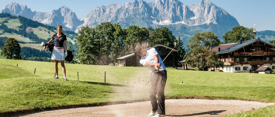 En golfspiller slår et slag fra et sandfang på en frodig bane med en bjergrig baggrund, der minder om Wagrain. En kvinde står i nærheden og ser under den klare blå himmel, mens træer indrammer en træbygning i baggrunden – en scene lige ud af en ideel sommerferie Østrig.