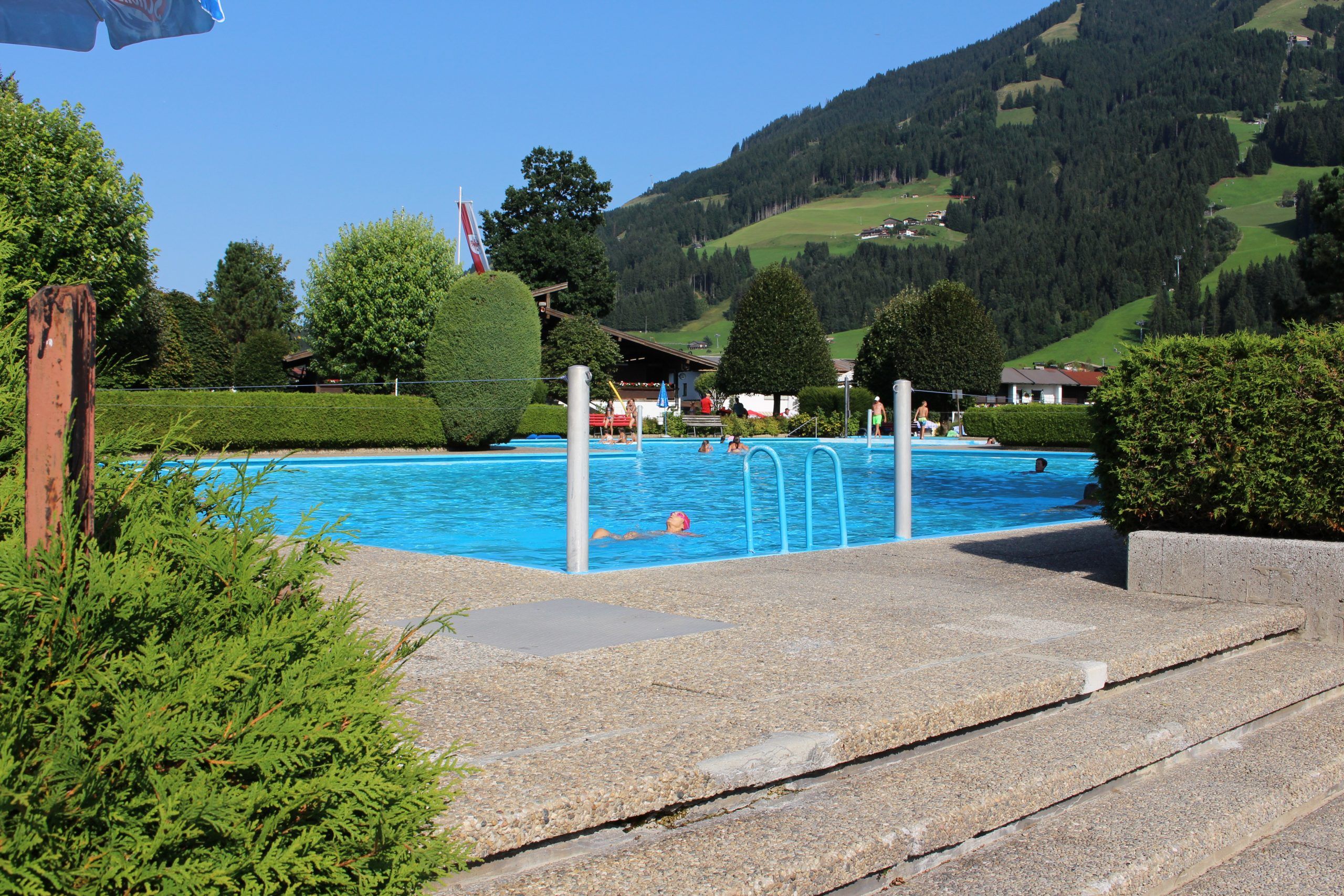 En fredfyldt udendørs swimmingpool, der minder om en Sommerferie i Westendorf, er omgivet af frodige grønne hække med mennesker, der svømmer. I baggrunden rejser sig en bjergskråning dækket af træer og pletter af åbent græs under en klar blå himmel.