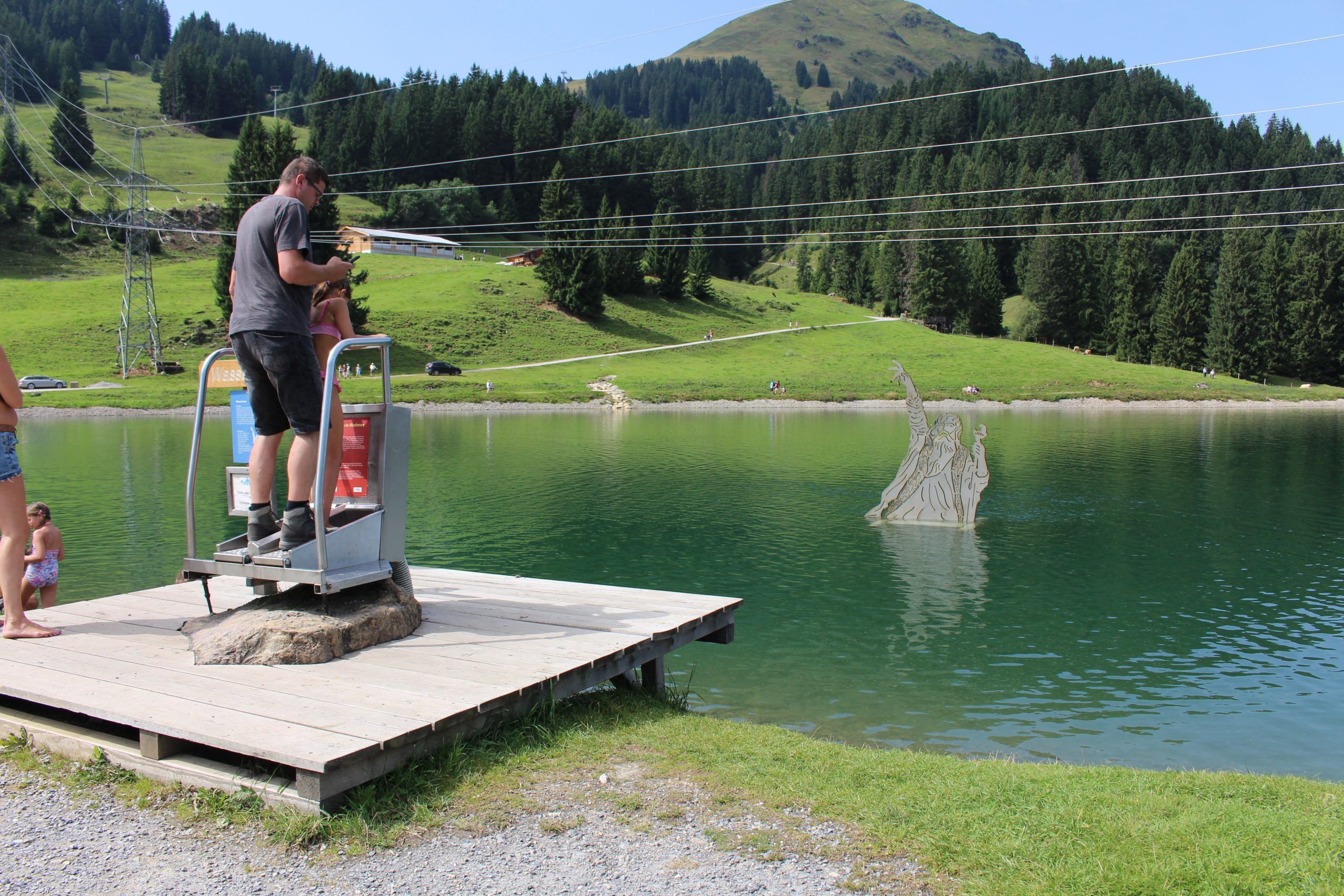 En man och ett barn står på en liten träbrygga vid en sjö i Westendorf och använder en myntdriven maskin. I sjön dyker en drakeskulptur upp ur vattnet, omgiven av gröna kullar och tallar – perfekt för sommarferie Österrig.